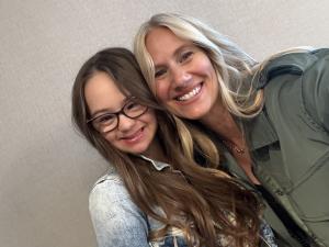 A smiling woman and a smiling girl take a close-up selfie together in front of a light-colored wall. The girl wears glasses and a denim jacket, and the woman wears a green jacket with long, wavy blonde hair. They are leaning in closely and appear happy an