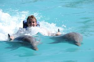 John “The Dolphin Expert” McNamara being pushed through bright blue water by two dolphins during a swim-with-dolphins experience.