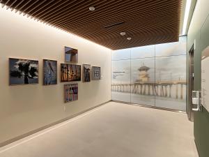 A quiet hospital corridor features a curated display of coastal-themed artwork on the left wall, including photographs of palm trees, a pelican, surfboards, and lifeguard towers. At the end of the hallway, a large wall graphic shows a soft, misty image of