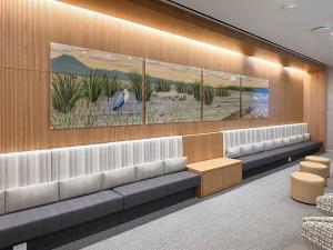 A long, softly lit seating area features a wood-paneled wall with a four-panel nature artwork depicting marsh grasses, birds, and distant mountains. Upholstered bench seating runs the length of the wall, accented by small wood tables and neutral-toned ott