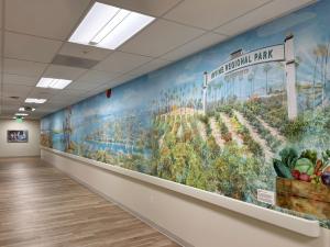 A long hospital corridor features a vibrant, panoramic mural celebrating Irvine Regional Park. The artwork depicts rolling orchards, palm-lined paths, a scenic valley landscape, and the park’s iconic entrance arch, with a crate of colorful produce in the