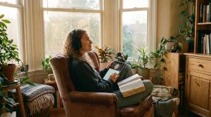 A young woman wearing headphones and holding a tablet displaying a glowing cross, sitting in a cozy, sunlit room filled with plants. She has an open physical Bible on her lap, illustrating the modern blend of technology and faith for daily devotion and me