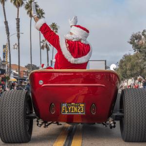Santa Claus in a red suit waves to the crowd while riding in a bright red classic hot rod car down a street lined with spectators at the La Jolla Christmas Parade.