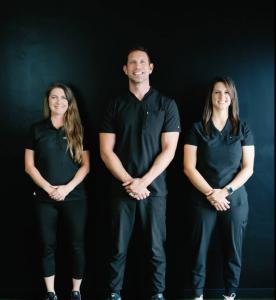 Three staff members from Accelerate: Sport and Spine Rehab standing in front of a black background. A man [Founder James R Jonas] stands in the center, with two women standing on his left [Carly Popst] and right [Alyssa Watts].