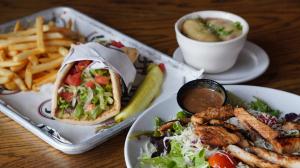 A tray with a gyro stuffed with lettuce, tomatoes, and meat served alongside French fries and a pickle spear, next to a grilled chicken salad with dressing and a small bowl of soup in the background.