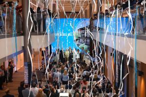 Grand opening ceremony in multi-level atrium with speakers on central stage, large crowd on multiple floors, and white and blue streamers hanging from ceiling.