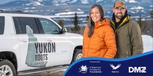 A smiling man and woman stand side by side outdoors in a snowy Yukon landscape, with mountains in the background. They are wearing winter jackets and standing next to a white SUV branded with the Yukon Roots Adventure Co. logo. A branded banner at the bot
