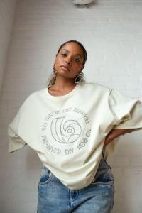 A woman with large hoop earrings and light-wash jeans wears a loose-fitting cream crewneck shirt. The shirt has a circular graphic design on the chest featuring a swirl pattern and the text "NO SINGLE USE FASHION" and "PRE-LOVED DIY HQB."