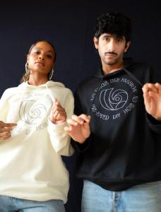 A man and a woman pose together wearing matching hoodies. The woman wears a cream hoodie, and the man wears a black one. Both hoodies feature a circular graphic design with text around it that says "NO SINGLE USE FASHION" and "PRE-LOVED DIY HQB."