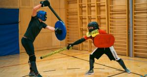 Two athletes wearing helmets and protective gear spar with padded sword and shield in an indoor sports hall.
