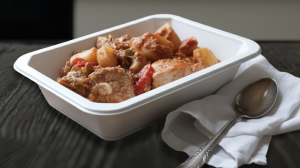 A serving of Chicken Stifado with Yukon potatoes in a white meal tray, placed on a dark wooden surface with a spoon and napkin beside it, with a blurred kitchen background.