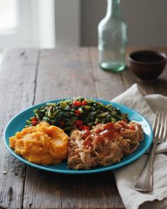 A plate of Memphis-style shredded BBQ pork topped with sauce, served alongside sweet potato mash and sautéed kale with red peppers on a rustic wooden table.