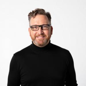 Professional headshot of Jason Riggs wearing a black turtleneck against a white background.