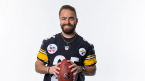 A man wearing a Pittsburgh Steelers jersey with a “Fan of the Year” patch smiles while holding a football against a plain white background.