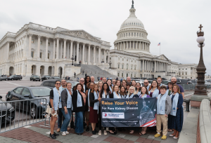 A large group of IgA Nephropathy Foundation advocates stand in front of the U.S. Capitol building holding a banner that reads “Raise Your Voice for Rare Kidney Disease.” The group includes patients, caregivers, staff, and supporters wearing light blue adv