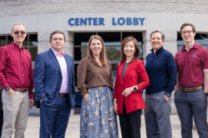 A group of six GSSM A.I. Faculty Fellows stand in front of a building entrance labeled “Center Lobby.” From left to right: Wilson Harvey, Spencer Tyce, Jennifer Brown, Haiqing Kaczkowski, Stephen Kaczkowski and Reggie Bain. Two additional fellows, Taylor