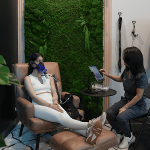 Patient seated in a recliner at MetaLab wearing a PNOĒ metabolic testing mask during an RMR test, while a clinician monitors breath analysis data on a tablet in a calm testing room.