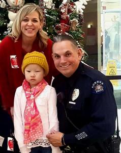 Founder Jim Seneca kneels beside a young child wearing a yellow knit hat and scarf, with a hospital staff member standing behind them near a decorated Christmas tree.