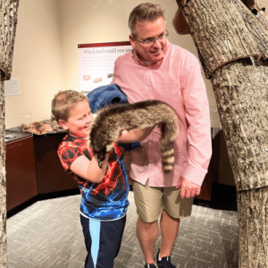 A child holds a raccoon while an adult stands next to them.