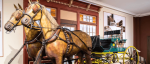 Two life-like, beige horses harnessed to a vintage carriage inside a museum, with historical photographs on the walls.