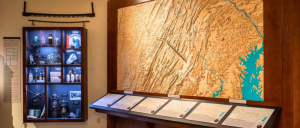 A museum display featuring a wooden relief map of Virginia, artifacts in a lit cabinet, and informational panels on a tabletop.