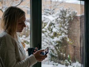 Woman experiencing seasonal depression looking at phone by snowy winter window, representing holiday mental health challenges treated by Montesano Psychological Center in Antioch Illinois