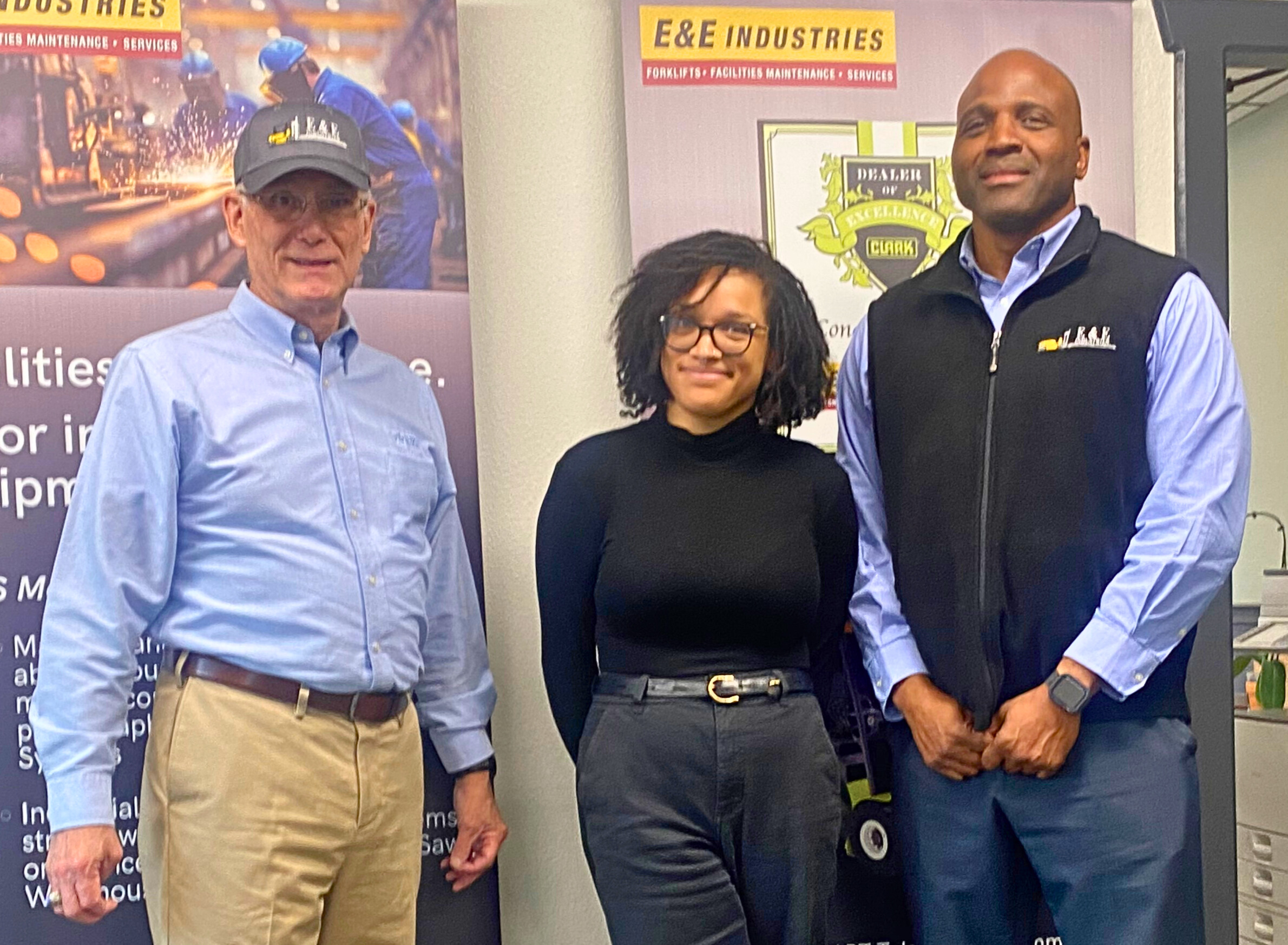 Chuck Moratz, Sharelle Harris and Fernando Harris are standing in front of the CLARK New Dealer of the Year banner at E&E's San Diego Office conference room.