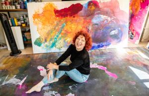 Artist Clara Berta sitting on the studio floor, smiling, with vibrant paintings around her.