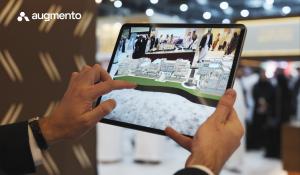 A person holds a tablet displaying an augmented reality model of a large real-estate development while standing at a busy event or exhibition. The AR experience overlays a 3D architectural visualization on the screen. The Augmento logo appears in the uppe