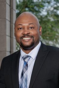 Professional Photgraph of Ernest Manning Jr standing with a black building in back ground and bushes.