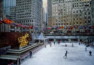 Rockefeller Center ice skating rink in New York City with skaters, holiday lights, international flags, and the golden Prometheus statue.