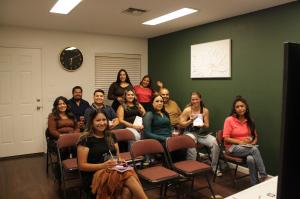 A group of people seated in rows of chairs inside a small meeting room, smiling toward the camera. Some attendees hold notebooks or drinks, while two women stand in the back posing and waving. The room has a green accent wall with a white floral artwork,