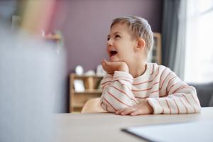 Smiling child in a striped sweater sits at a table, chin resting on hand, looking off-camera during a playful learning session in a bright therapy room.