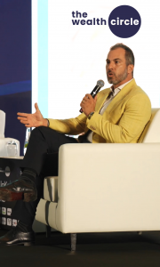 Nick Ayala speaking on stage at a business event in Dubai, UAE, holding a microphone, with The Wealth Circle logo displayed above him.