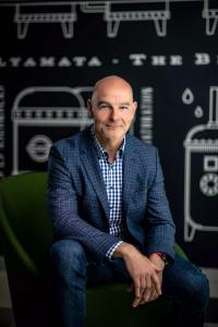 Portrait of Gabor Bekefi, an experienced global executive, seated and smiling in a business-casual jacket against a dark background with industrial illustrations.