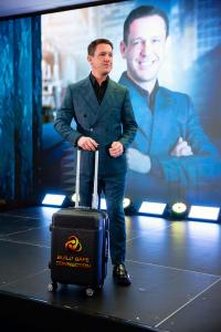 Rony Jabour standing on stage at the Build Safe Connection 2025 Conference in Boston, holding a branded BSC suitcase with his portrait displayed on the screen behind him.