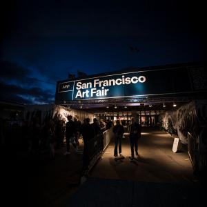 Night-time view of the San Francisco Art Fair entrance with crowds walking into the venue under a brightly lit sign.