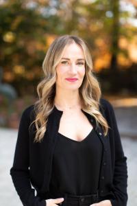 Professional headshot of a business executive standing outdoors, wearing a black top and blazer, with wavy blonde hair and a confident expression.