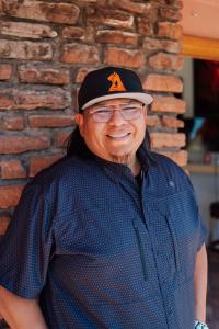 Portrait of Navajo photographer Lionel Bigthumb wearing a black patterned shirt and branded hat, standing and smiling against a rustic brick wall.