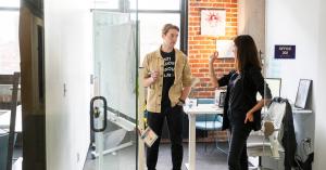Two people talking inside a glass-walled office at Elevator’s Omaha location, with a whiteboard, desk setup, and exposed brick wall visible.