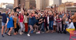 A large group of people smiling and posing together on Elevator’s rooftop deck in Lincoln, with downtown buildings in the background.