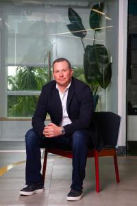 Andrew Beachler, CEO of Beachler Investments, is seated in a modern South Florida business setting with palm trees visible through glass behind him. He leads Florida real estate redevelopment initiatives including mixed-use development, adaptive reuse, an