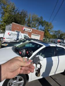 Hand holding three Toyota remote keys and a key fob in front of a white car parked outside Dan's Lock and Key in Bristol, PA, with the company van and building in the background.