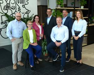 Seven Karrikins Group team members pose for a professional group photo in a modern office with plants and abstract line art, with two seated on stools in front and five standing behind, all in business attire smiling at the camera, from left to right Dani