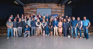 Large group of diverse real estate professionals standing together and smiling on an indoor stage at the Real Estate Forum (REF) Leander x Austin Chapter event, with a wood-paneled wall and projector screen in the background.