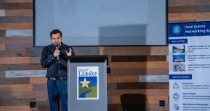 Ravi Polishetty, founder of Real Estate Forum (REF), stands at a City of Leander podium speaking into a microphone at the REF Leander x Austin Chapter event, with a presentation screen and REF networking banner in the background.