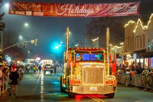 A large semi truck covered in lights driving under a holiday banner