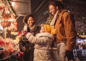Happy family of three browsing holiday treats and decorations at an outdoor market, with child in yellow knit hat smiling between two parents.