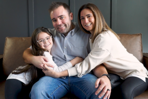 Blood clot survivor Dan Walsh sits on a couch smiling with his wife and young daughter.