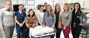 A group of healthcare professionals pose together in a medical setting, with medical equipment and a bed visible in the background.
